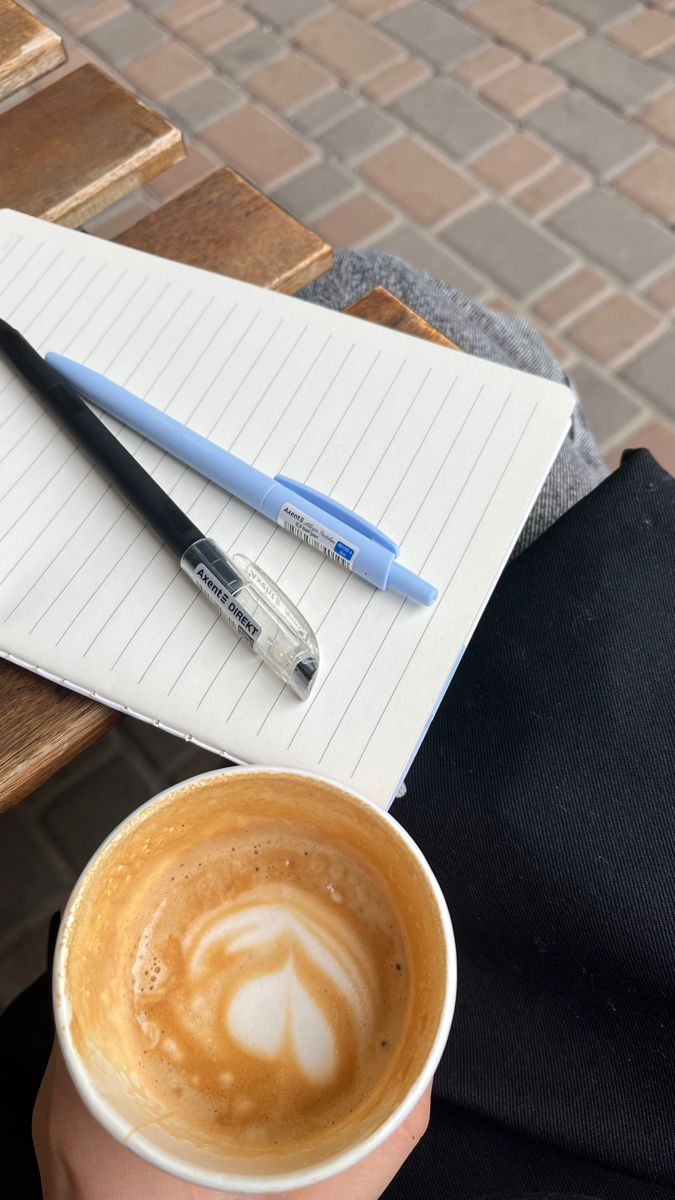 Caderno aberto com canetas e um café com latte art sobre as pernas